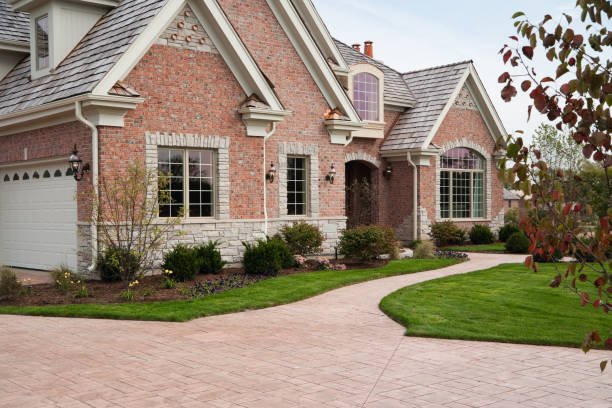 A brick driveway made of stamped concrete leads to a large, elegant house surrounded by greenery.
