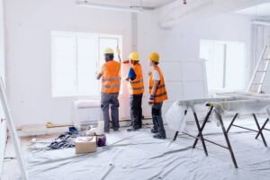 Three men in orange vests and hard hats repair a chipped concrete surface in a room