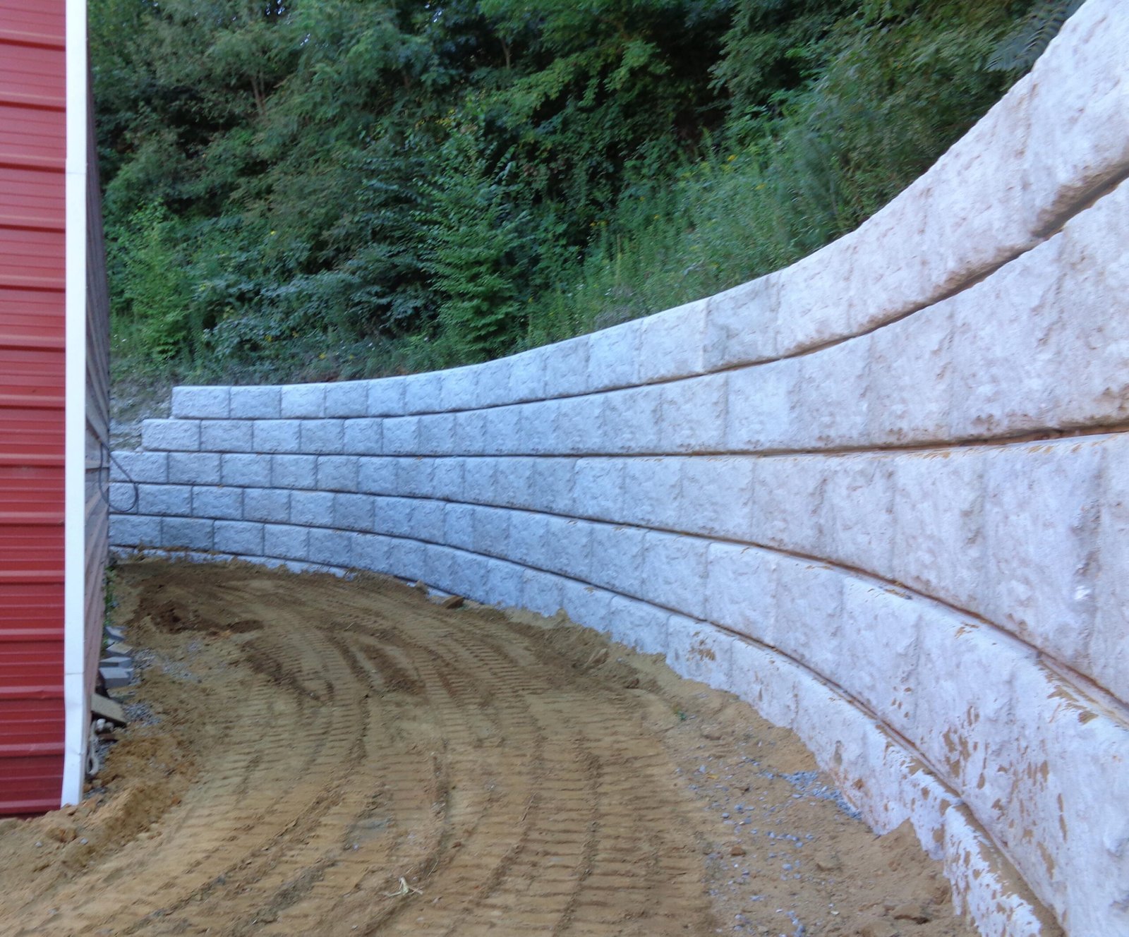 A curved concrete retaining wall bordering a dirt path next to a red metal building and a wooded hillside.